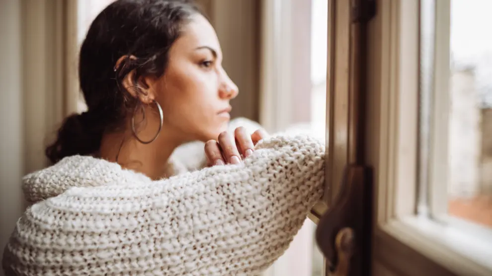 Mujer pensativa y seria frente a una ventana.